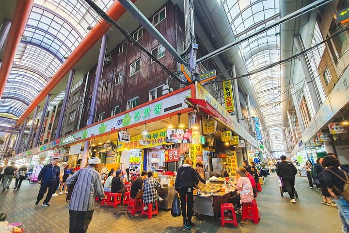 Bustling traditional market at the start of the Busan Food Crawl, filled with vibrant food stalls, red stools, and locals enjoying street snacks under colorful signs and a towering arched ceiling.
